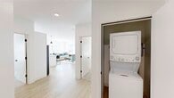 This well-appointed single-floor residence offers two bedrooms and two bathrooms within a thoughtfully designed, open layout. The interior is defined by a clean, modern aesthetic, with white walls, light wood-style flooring in the main living areas, and black hardware accents throughout that add a contemporary contrast.

The kitchen is a standout feature, equipped with white shaker cabinetry, stainless steel appliances including a range and built-in microwave, and a large white island with pendant lighting above—ideal for dining and entertaining. Natural light filters in through multiple windows fitted with blinds, brightening both the kitchen and the adjacent living area, which offers generous room for seating arrangements.

The primary bedroom is notably spacious, featuring plush carpeting, large windows that invite natural light, and a sliding-door closet. The second bedroom is similarly appointed with carpet flooring and ample natural light. Both bathrooms feature wood-tone flooring, white vanities with black hardware, and shower setups with clean, minimal finishes.

A dedicated in-unit laundry closet with a stacked washer and dryer adds everyday convenience. The overall ambiance is bright, modern, and functional throughout.