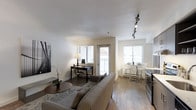 This single-floor space showcases a thoughtfully designed open-concept layout that combines the living, dining, and workspace areas into one cohesive environment. The living area is anchored by a comfortable grey sofa and a round coffee table, complemented by hardwood flooring that runs throughout, adding warmth and continuity to the space.

Natural light is a defining feature, with multiple windows fitted with white horizontal blinds allowing for adjustable brightness. A glass-paneled door leads to a small outdoor balcony, further enhancing the sense of openness. The dining area features a round white table paired with modern acrylic chairs, creating an airy, contemporary feel. Adjacent to it, a sleek kitchen with dark cabinetry provides a functional contrast to the light tones throughout.

A dedicated desk area near the balcony door offers a practical workspace without disrupting the flow of the room. Overhead, a track lighting fixture provides targeted illumination, while a table lamp adds a softer ambient glow. Subtle decor elements, including wall art and a small orchid plant, contribute to the clean, minimalist aesthetic of the overall space.