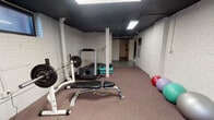 Located at 34 Hill Street in Morristown, NJ, this dedicated fitness facility offers a comprehensive, single-floor workout environment designed to accommodate a wide range of exercise disciplines. The space is characterized by its white-painted brick walls, dark ceiling with exposed structural elements, and durable carpeted flooring throughout, creating a functional and focused atmosphere.

The main workout area is generously proportioned and well-lit by recessed fluorescent panel lighting. A full wall of mirrors along one side enhances the sense of openness and allows for form-checking during exercise. Cardio equipment includes a treadmill and an arc trainer elliptical, while strength training is supported by a multi-station cable machine, a barbell rack with weight plates, and a flat/adjustable workout bench. A tiered dumbbell rack stocked with a comprehensive range of free weights is positioned along the mirrored wall for easy access.

A secondary section of the space provides a more open area ideal for stretching and floor-based workouts, featuring a collection of stability and exercise balls in varying sizes, stacked exercise mats, and additional floor space. Small ground-level windows along the upper walls allow in natural light and provide ventilation. The overall layout is practical and efficient, making this space well-suited for individual training sessions or small group fitness use.