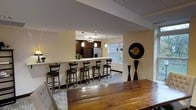 This well-appointed single-floor space is thoughtfully designed for both casual gatherings and more structured entertaining. At the heart of the layout is an open-concept kitchen and bar area, separated from the main gathering space by a sleek white countertop that doubles as a serving surface. A row of dark wood swivel bar stools lines the counter, providing ample seating for guests while maintaining a clear sightline into the kitchen. The kitchen itself is outfitted with dark espresso cabinetry, a stainless steel refrigerator, and elegant pendant lighting that hangs at varying heights, adding a layered, modern touch to the space. Natural light filters in through a window adjacent to the kitchen, brightening the area with a warm, welcoming glow. The main gathering area features a substantial rustic wood table in the foreground, contrasting beautifully with the more polished finishes throughout. A decorative oversized clock and sculptural floor accents add artistic character to the space. Neutral wall tones and a patterned area rug tie the different zones together, creating a cohesive and inviting atmosphere. Overall, the space strikes a balance between functional design and stylish décor, making it well-suited for community events, receptions, or social gatherings.