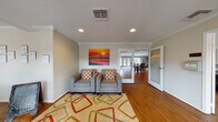 This single-floor community space is thoughtfully designed with a coastal aesthetic that carries throughout every area. The entrance opens into a comfortable lounge featuring warm hardwood-style flooring, crown molding, recessed lighting, and a pair of grey armchairs arranged atop a vibrant geometric area rug. A bold sunset beach painting anchors the wall, setting an inviting tone from the moment you arrive. A chalkboard easel and framed wall art add a welcoming, communal character to the entry area. Moving deeper into the space, an expansive open-plan great room unfolds, bathed in natural light from large windows and glass French doors. Soft teal-painted walls complement the rich hardwood floors throughout. This area accommodates a spacious lounge with a fireplace, wall-mounted television, and comfortable seating, alongside a dedicated dining area with a full table and chairs. A kitchen bar with granite countertops and pendant lighting in amber tones provides additional seating. Elegant frosted glass panel doors featuring nautical motifs offer a distinctive architectural detail. The fitness center is well-equipped with cardio machines, strength training equipment, a medicine ball rack, and a large wall mirror, with direct access to an outdoor pool area visible through oversized windows.