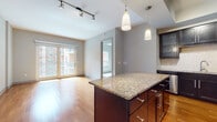 This well-appointed single-floor residence offers a thoughtfully designed layout that balances modern aesthetics with functional living. The open-concept kitchen serves as the heart of the space, showcasing rich dark cabinetry with sleek brushed-nickel hardware, a striking granite-topped island with pendant lighting overhead, a classic tile backsplash, and a full suite of stainless steel appliances including a refrigerator, range, microwave, and dishwasher. A tray ceiling with recessed lighting adds architectural interest above the kitchen area. Warm hardwood flooring flows throughout the main living area, which is bathed in natural light from glass French doors that open to an outdoor view of the surrounding urban landscape. Track lighting provides additional illumination throughout the main living space. Two bedrooms, each carpeted for comfort, feature large windows that invite natural light and offer views of the surrounding cityscape, with ceiling fans providing year-round climate control. The unit includes two bathrooms: one featuring a soaking tub with a tiled surround and a dark vanity with granite countertops, and a second bathroom with a generous walk-in closet, tiled flooring, and matching dark cabinetry. The cohesive design elements — granite surfaces, dark wood tones, and neutral wall colors — lend the space a polished, contemporary character throughout.