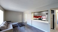 This single-floor space offers a clean, modern aesthetic defined by an open-concept layout that seamlessly connects the living area to the kitchen. The living space is bright and functional, featuring smooth grey hardwood-style flooring that extends throughout, neutral grey walls, and a textured ceiling. A minimalist sofa and coffee table arrangement anchors the room, while a doorway leads to an adjoining space, adding to the unit's overall flow and accessibility.

The kitchen is a standout feature, framed by a peninsula with a crisp white countertop that doubles as a casual dining or prep surface. Dark charcoal cabinetry with a linear tile accent panel creates a sleek backdrop, while a bold red subway tile backsplash adds a striking pop of color and visual interest. The kitchen is equipped with a stainless steel electric range, an integrated range hood, and a single-basin sink, providing all essential cooking and prep amenities.

The interplay between the dark cabinetry, vivid backsplash, and light countertops creates a dynamic contrast that gives the kitchen a contemporary, well-designed feel. The open sightlines between the kitchen and living area make the space feel airy and connected, ideal for everyday living.