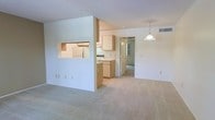 This single-floor space features an open-concept layout that connects the living area to the kitchen, creating a sense of spaciousness and ease of movement. The living area is finished with wall-to-wall neutral carpeting, contributing to a clean and understated aesthetic, while white walls and a textured ceiling provide a bright, airy backdrop throughout.

The kitchen is partially separated by a half-wall with a pass-through window, maintaining visual connectivity between the cooking and living spaces. Light wood cabinetry lines the kitchen walls, offering ample storage, and standard appliances are included. The design allows natural light to filter through the space, keeping the kitchen bright and functional.

A pendant light fixture hangs above the dining or transitional area, serving as a subtle focal point. A hallway visible from the main living space leads to additional rooms beyond, suggesting a practical and well-organized floor plan. HVAC vents are integrated into the ceiling, ensuring climate control throughout. Overall, the space presents a straightforward, functional layout with neutral finishes suited for a variety of furnishing styles.