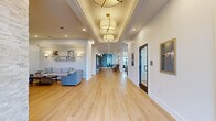 This thoughtfully designed single-floor amenity space offers a diverse range of functions, seamlessly blending wellness, recreation, and social gathering areas within one expansive layout. The entry foyer welcomes visitors with light-washed hardwood floors, floor-to-ceiling black-framed glass doors, and curated accent seating that establishes a refined, contemporary tone.

The main corridor extends the full length of the facility, featuring warm hardwood flooring, coffered ceilings with layered pendant lighting, recessed illumination, and integrated wall sconces. Seating alcoves line the hallway, offering intimate areas for conversation.

A central social lounge incorporates a stacked-stone structural column, herringbone-patterned accent wall, and coordinated dining and lounge seating. An adjacent kitchenette provides a white island with bar seating, teal tile backsplash, open shelving, and full appliances. A dedicated billiards room with a tray ceiling, globe chandelier, and textured accent wall adds a recreational focal point.

The expansive fitness center is divided into distinct zones: a cardio area lined with treadmills and ellipticals facing large windows with outdoor views, and a strength training zone equipped with cable machines, free weights, and resistance equipment on durable rubber flooring. Throughout, the space maintains a bright, upscale aesthetic defined by clean lines and carefully curated décor.