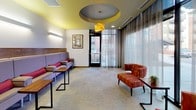 This contemporary lobby lounge is thoughtfully designed to offer a welcoming and comfortable environment. The space is anchored by a long, upholstered built-in bench with red accent pillows, set against a warm wood-paneled feature wall and illuminated by sleek wall-mounted linear sconces. Lightweight side tables with black metal frames are positioned conveniently throughout for guests' use.

A striking architectural ceiling detail — a layered circular tray with a bold yellow accent — draws the eye upward and serves as the focal point of the room, complemented by a sculptural yellow pendant light that adds a warm, artistic touch. The yellow ceiling color is carried throughout, creating a cohesive and energetic design palette.

Floor-to-ceiling sheer curtains line the expansive glazed exterior wall, softening the abundant natural light while maintaining views of the surrounding outdoor environment. Additional seating is provided by richly upholstered red patterned armchairs arranged in conversational groupings, adding warmth and texture to the neutral carpeted floor.

A framed art piece adorns one wall, contributing to the curated aesthetic, while a glass-panel entry door offers a clear sightline to the outdoors. Decorative etched-glass interior doors are visible further down the corridor, hinting at additional spaces beyond. The overall ambiance is polished, inviting, and well-appointed.
