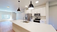 Located at 4912 Dunham Dr in Olympia, WA, this two-story home offers a thoughtfully designed layout spread across two floors. The main level, which comprises the larger portion of the living space, features an open-concept kitchen and living area finished with light hardwood flooring throughout. The kitchen is appointed with crisp white cabinetry, a large island with an undermount sink, stainless steel appliances including a French door refrigerator, electric range, and built-in microwave, all illuminated by stylish pendant lighting and recessed fixtures. A dedicated entrance leads to an open staircase with wood-tread steps and clean white railings, connecting both levels. The upper floor houses two bedrooms, including a spacious primary bedroom with vaulted ceilings, a ceiling fan, a large window allowing ample natural light, and direct access to a dedicated closet. The shared bathroom features a white vanity and is adjacent to a balcony offering views of surrounding mature trees. Neutral wall tones and consistent hardwood flooring create a cohesive, modern aesthetic throughout the home.