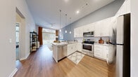 This thoughtfully designed single-floor residence offers a seamless blend of modern aesthetics and functional living. The open-concept kitchen and living area forms the heart of the home, highlighted by soaring vaulted ceilings and an abundance of natural light streaming through large windows. The kitchen is outfitted with crisp white shaker cabinetry, granite countertops, a peninsula island with a built-in sink, and a full suite of stainless steel appliances including a range, over-the-range microwave, dishwasher, and refrigerator. Stylish pendant lighting anchors the island, while wood-look flooring flows throughout the main living areas, adding warmth and continuity. The living area extends toward a large window with access to an outdoor space, enhanced by a ceiling fan overhead. The primary bedroom is a serene retreat, featuring plush carpet, tall ceilings, and generous windows that invite natural light. It connects to a large walk-in closet equipped with multi-tier wire shelving and hanging space. A secondary bedroom offers equal comfort with soft carpet and easy hallway access. The bathroom is finished with granite countertops, white cabinetry, wood-look flooring, and a tub-shower combination, completing this well-appointed residence.