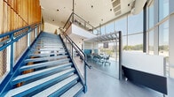 Located at 505 E St Louis St in the heart of downtown Springfield, Missouri, this contemporary two-floor commercial space offers a well-appointed environment designed for productivity and collaboration. The main floor greets visitors with large-format tile flooring, clean white walls, and an abundance of natural light streaming through expansive floor-to-ceiling glass curtain walls that frame striking panoramic views of the city skyline. A bold steel staircase with mesh railings serves as both a functional and architectural focal point, flanked by a dramatic vertical wood slat accent wall that adds warmth and texture to the modern interior. Seating on this level includes semi-private booth-style seating and individual lounge chairs, complemented by a sleek linear fireplace set into a tile-clad surround. The upper floor offers a more intimate setting, carpeted throughout and bathed in natural light from continuous glass walls and windows overlooking the downtown streetscape. Plush lounge chairs and compact side tables create relaxed gathering nodes, while private pod-style workstations provide focused work areas. Wide corridors with patterned carpet tile connect to additional adjacent spaces via elevator access. Throughout both floors, pendant and cylinder lighting fixtures, along with recessed ceiling elements, maintain a polished and cohesive modern aesthetic.