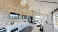 This single-story clubhouse-style space is thoughtfully designed to offer a variety of functional and comfortable gathering areas. The main lounge area serves as the heart of the space, featuring a striking floor-to-ceiling stacked stone fireplace with a rustic wood mantel, plush seating arrangements, and carpeted flooring. French doors with bold black frames flank the fireplace, filling the room with natural light and providing direct access to an outdoor area.

Adjacent to the lounge, a formal seating area is defined by elegant cream upholstered furniture, soft accent pillows in muted tones, and refined chandelier lighting. Black-trimmed glass-panel doors connect this zone to neighboring spaces while maintaining an open, airy feel.

The kitchenette area is particularly notable, featuring soaring vaulted ceilings clad in warm exposed brick, light gray cabinetry with a clean white countertop, a built-in sink, and stainless steel appliances. Oversized drum pendant lights hang overhead, adding a modern touch. Large windows at the end of this area bring in ample natural light and offer exterior views.

A welcoming entry foyer with tall ceilings, a wood-and-glass front door, chandelier lighting, and tile flooring provides an inviting transition into the interior. Throughout, the design balances contemporary finishes with warm, textural elements.