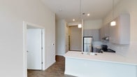 This well-appointed 2-bedroom, 2-bathroom residence is designed with a modern, open-concept layout that maximizes both space and functionality. The kitchen serves as the heart of the home, featuring sleek gray cabinetry, a classic subway tile backsplash, stainless steel appliances including a refrigerator and range, and a spacious white countertop island with an integrated sink. A stylish pendant light hangs above the island, adding a refined touch to the space. The kitchen flows seamlessly into the main living area, creating an inviting environment ideal for both everyday living and entertaining. Rich, dark hardwood-style flooring runs throughout, providing a warm contrast to the crisp white walls and ceilings. Elevated ceilings contribute to an airy, open ambiance, while recessed lighting ensures the space is well-lit throughout. A smart thermostat is conveniently installed on the wall, reflecting the home's thoughtful, modern amenities. The layout also includes well-defined hallways leading to the two bedrooms and bathrooms, offering a practical and comfortable flow. Overall, this residence combines contemporary design with functional living across a single, cohesive floor plan.