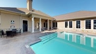 This single-story clubhouse serves as the centerpiece of a well-maintained residential community, offering a range of amenities designed for comfort and leisure. The building features a classic architectural style characterized by a neutral stucco exterior, stone veneer accents along the base, and a traditional pitched roof with asphalt shingles. Large multi-pane windows and elegant French doors flood the interior with natural light while providing views of the surrounding outdoor spaces.

The covered patio entry is framed by stately white columns and finished with a natural stone tile floor, creating a welcoming transition between the interior and the pool area. The outdoor space centers around a generously sized, crystal-clear swimming pool with a dedicated entry staircase, surrounded by a spacious concrete pool deck ideal for lounging. An adjacent spa or wading area adds to the recreational appeal.

Outdoor seating and lounge areas are thoughtfully arranged around the pool deck, complemented by mature trees and lush landscaping. A wrought iron perimeter fence encloses the pool area, ensuring privacy and safety. The backdrop of multi-story residential buildings with balconies provides context for the community setting. Overall, the space presents a polished, resort-inspired atmosphere suited for relaxation and social gathering.
