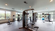 This fitness center is a comprehensive, single-floor facility designed to support a full range of workout routines. The main exercise area is spacious and open, featuring durable rubber flooring and a lineup of modern cardio machines, including Matrix treadmills and elliptical trainers, positioned along expansive floor-to-ceiling windows. These large, black-framed windows flood the space with natural light and offer unobstructed views of the surrounding greenery and outdoor environment, creating an energizing atmosphere. Strength training is well-represented with a multi-station cable machine system, a dedicated free weights bench, and stability balls for functional training. Wall-mounted flat-screen televisions are positioned throughout for entertainment during workouts. A separate, distinct cycling room is highlighted by a bold, colorful motivational mural reading "STRONGER," adding an energetic aesthetic. This room includes a Matrix spin bike, a built-in storage and counter area with yellow cabinetry, and a window providing additional natural light. The entrance area includes an accessible dual-level water fountain and restroom access, ensuring convenience and accessibility. Recessed lighting throughout the facility maintains a clean, modern ambiance, making the space both functional and visually inviting.