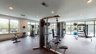 This fitness center is a comprehensive, single-floor facility designed to support a full range of workout routines. The main exercise area is spacious and open, featuring durable rubber flooring and a lineup of modern cardio machines, including Matrix treadmills and elliptical trainers, positioned along expansive floor-to-ceiling windows. These large, black-framed windows flood the space with natural light and offer unobstructed views of the surrounding greenery and outdoor environment, creating an energizing atmosphere. Strength training is well-represented with a multi-station cable machine system, a dedicated free weights bench, and stability balls for functional training. Wall-mounted flat-screen televisions are positioned throughout for entertainment during workouts. A separate, distinct cycling room is highlighted by a bold, colorful motivational mural reading "STRONGER," adding an energetic aesthetic. This room includes a Matrix spin bike, a built-in storage and counter area with yellow cabinetry, and a window providing additional natural light. The entrance area includes an accessible dual-level water fountain and restroom access, ensuring convenience and accessibility. Recessed lighting throughout the facility maintains a clean, modern ambiance, making the space both functional and visually inviting.