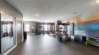 Located at 2101 Railway Ln. in Marysville, Ohio, this fitness center is a comprehensively outfitted exercise facility designed to support a wide range of workout styles and fitness goals. The space is finished with durable dark rubber flooring throughout, providing both safety and functionality for high-intensity training.

At the heart of the facility stands a professional-grade strength training area featuring dual power racks with red and black framing, loaded with pull-up bars, weight plate storage, and cable attachments. A full dumbbell rack and an assortment of medicine balls, kettlebells, and weighted plates round out the free weight offerings. Additional cardio and functional training equipment, including step platforms and stability balls, provide versatility for varied workout routines.

A striking large-scale abstract mural in vivid blues, golds, and warm tones serves as a bold focal point along one wall, adding an energizing artistic element to the space. Full-length mirrors are strategically placed to support form checks during exercise.

The facility also includes a wooden sauna unit, offering a recovery option alongside the workout areas. Separate restroom facilities are accessible via warm-toned wooden doors along the main wall. Recessed lighting and windows with natural light contribute to a bright, motivating atmosphere throughout this well-appointed fitness space.