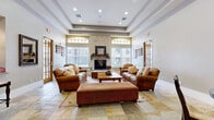 This single-floor community space is thoughtfully designed to foster comfort and social interaction. The centerpiece of the space is a spacious lounge area anchored by a striking floor-to-ceiling stone fireplace, flanked by arched transom windows that invite abundant natural light. A tray ceiling with recessed lighting adds architectural depth, while warm-toned natural stone tile flooring flows seamlessly throughout the entire space, creating a cohesive and inviting ambiance. Plush leather sofas and upholstered seating are arranged around a wooden coffee table, complemented by a large ottoman and a patterned area rug, offering ample room for relaxation and gathering. French-paned doors with warm wood frames provide easy access and enhance the open, airy feel of the lounge. Adjacent to the main seating area, a casual dining nook features a round glass-top table with cushioned wooden chairs beneath colorful wall art, creating an intimate setting for smaller gatherings. The well-appointed kitchen is equipped with light maple cabinetry, granite countertops, a tile mosaic backsplash, and stainless steel appliances including a gas range and built-in microwave, making it fully functional for entertaining. Together, these spaces form a warm and versatile community environment.