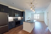 This single-floor unit showcases a thoughtfully designed open-concept layout where the kitchen flows seamlessly into the living area. The kitchen is a standout feature, outfitted with floor-to-ceiling dark navy shaker-style cabinetry accented by gold bar pulls, dark granite countertops, and a full suite of stainless steel appliances including a refrigerator, electric range, and over-the-range microwave. A herringbone-patterned tile backsplash adds a refined decorative touch behind the range. A central kitchen island with a white base, dark granite surface, and a gold-toned faucet with an undermount sink serves as the focal point of the space, illuminated by a cluster of elegant gold pendant lights overhead.

The open living area extends from the kitchen, featuring warm wood-style plank flooring, white walls, and a ceiling fan. Natural light enters through large windows with horizontal blinds, offering views of the surrounding urban environment. The bedroom is carpeted and similarly benefits from a generously sized window with outdoor views.

The bathroom is cleanly finished with large-format neutral tile surrounding a soaking tub and shower combination, complemented by gold and matte black fixtures, a dark-vanity with granite countertop, and light tile flooring throughout.
