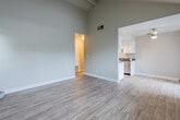 This single-story residence is defined by its dramatic vaulted ceilings with exposed beams, which lend an open, airy quality to the main living area. Light wood-style flooring runs throughout the living spaces, complementing the neutral gray wall tones and clean white baseboards to create a cohesive, modern aesthetic.

The living area is generously proportioned, with multiple windows fitted with horizontal blinds allowing natural light to filter in. The space flows openly toward the kitchen, which is laid out in a galley configuration. The kitchen features white shaker-style cabinetry on both sides, dark countertops, a stainless steel sink, gas range, and dishwasher, along with a skylight overhead that brightens the space considerably.

The bedroom offers a quieter atmosphere, finished with plush neutral carpeting and a large window with views of surrounding greenery, providing ample natural light. The full bathroom includes a tub and shower combination, a white vanity with dark countertop, a large mirror, pendant lighting, and wood-style flooring, offering a clean and functional design. The overall ambiance throughout is neutral, bright, and move-in ready.