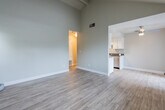This single-story residence is defined by its dramatic vaulted ceilings with exposed beams, which lend an open, airy quality to the main living area. Light wood-style flooring runs throughout the living spaces, complementing the neutral gray wall tones and clean white baseboards to create a cohesive, modern aesthetic.

The living area is generously proportioned, with multiple windows fitted with horizontal blinds allowing natural light to filter in. The space flows openly toward the kitchen, which is laid out in a galley configuration. The kitchen features white shaker-style cabinetry on both sides, dark countertops, a stainless steel sink, gas range, and dishwasher, along with a skylight overhead that brightens the space considerably.

The bedroom offers a quieter atmosphere, finished with plush neutral carpeting and a large window with views of surrounding greenery, providing ample natural light. The full bathroom includes a tub and shower combination, a white vanity with dark countertop, a large mirror, pendant lighting, and wood-style flooring, offering a clean and functional design. The overall ambiance throughout is neutral, bright, and move-in ready.