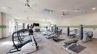 Located at 5776 Thomaston Rd in Macon, Georgia, this fitness center occupies a single floor and offers a generously sized, open workout environment. The space is designed with functionality and comfort in mind, featuring light-toned carpet flooring, white walls accented by a distinctive teal and yellow horizontal stripe that runs along the perimeter, and a bright, airy atmosphere created by multiple large windows that allow abundant natural daylight to fill the room.

The ceiling is outfitted with recessed lighting, ventilation, and several ceiling fans that ensure comfortable air circulation throughout the space. A wall-mounted flat-screen television provides entertainment for users during workouts.

The fitness floor is comprehensively equipped to support a full range of training styles. Cardio equipment includes treadmills and elliptical cross-trainers, while the strength training section features multiple cable and selectorized weight machines suited for various muscle groups. A dedicated free weights area offers a full dumbbell rack alongside kettlebells and weight benches. Additional functional fitness accessories, including stability balls and medicine balls on a wall-mounted rack, round out the facility's offerings. The overall ambiance is clean, modern, and well-organized, providing an inviting environment for a complete workout experience.