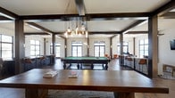 The Colony's amenity lounge is a thoughtfully designed, single-floor gathering space that blends rustic and contemporary elements throughout its open layout. The room is anchored by a full-sized billiard table positioned at the center, illuminated by an eye-catching cluster of Edison bulb pendant lights suspended from a structural beam by industrial chains. Dark wood beams frame the ceiling, adding architectural character and a warm contrast to the bright white walls and ceiling above.

Large windows along the perimeter flood the space with natural light while offering views of the surrounding exterior. The hardwood flooring ties the room together, complementing the rich tones of the wood furnishings, including a substantial communal table in the foreground. High-top tables and bar-style seating are arranged around the edges of the room, creating versatile areas for socializing or casual work. A seating lounge with upholstered chairs is visible in the background, providing a relaxed retreat. Wall sconces contribute to the layered lighting scheme, enhancing the inviting and social ambiance of the space.