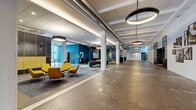 This expansive single-floor commercial space masterfully blends industrial heritage with contemporary design. Polished concrete floors stretch the full length of the open floor plan, reflecting light and reinforcing a sense of generous, uninterrupted space. White-painted wood-plank ceilings with exposed structural beams and visible HVAC ductwork lend an authentic industrial character, while a series of striking circular pendant lights illuminate the corridors and open areas with warm, focused light.

The space is anchored by bold design choices throughout. A vibrant teal accent wall adorned with a vintage bicycle mural and large painted numerals adds a dynamic focal point, complementing the retro-inspired yellow lounge chairs and dark leather seating that define casual gathering areas. Steel-framed grid partition doors and windows provide flexible room division while maintaining a cohesive industrial-modern aesthetic.

A long central corridor connects distinct zones, flanked by black steel-framed interior windows and original brick-textured wall features that nod to the building's history. Gallery-style photo displays line portions of the walls, adding a curated, creative atmosphere. A lounge area with built-in cabinetry, a television, and accent seating offers a polished, functional retreat. Double glass entry doors draw in natural light at one end, completing the bright, airy ambiance of this versatile and characterful commercial space.