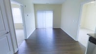 This single-floor space presents a clean, functional layout highlighted by rich, dark hardwood flooring that extends throughout the main living area, offering a sleek contrast against the light-toned walls. The room is generously proportioned, providing an open and airy feel well-suited for a variety of furniture arrangements. A prominent set of sliding glass doors serves as a focal point along the far wall, fitted with white vertical blinds that allow for adjustable natural light control. When opened, these doors likely provide access to an outdoor area, further enhancing the sense of openness. The walls are finished in a neutral, soft yellow-white tone that complements the flooring and contributes to a bright overall atmosphere. White baseboards run cleanly along the perimeter, adding a crisp, finished look. A doorway visible to the left suggests connectivity to additional rooms or areas within the unit. The space transitions from carpet near the entryway to hardwood in the main area, subtly defining zones within the open layout. Overall, the space offers a straightforward, well-lit environment with practical finishes.