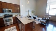 This single-floor residence offers an open-concept design that seamlessly integrates the kitchen, dining, and living areas into one cohesive and functional space. The kitchen is anchored by a substantial central island topped with speckled granite countertops, providing ample prep space and casual seating. Rich, dark wood cabinetry lines the walls, offering generous storage, while a stainless steel sink and modern pendant lighting add refined finishing touches. The warm-toned hardwood flooring flows continuously throughout, lending a sense of unity and warmth to the entire space. Notably high ceilings amplify the sense of openness, creating an airy atmosphere that feels both expansive and inviting. The living area is thoughtfully arranged with comfortable seating, complemented by carefully chosen décor including a large abstract artwork that adds a pop of color and personality. A round dining table with woven accent chairs creates a distinct yet connected dining zone. The neutral, light-toned walls serve as a versatile backdrop, allowing the natural and ambient lighting to enhance the overall brightness of the space. Floor lamps positioned throughout provide layered lighting options, making this residence as practical as it is visually appealing.