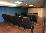 Located at 3901 Indian School Rd NE, this purpose-built screening room delivers an authentic cinema experience in a compact, well-appointed setting. The room is anchored by a large wall-mounted projection screen positioned at the front, paired with a ceiling-mounted projector that provides a sharp, wide viewing angle for all seats. Rows of dark upholstered theater-style seats are arranged on a tiered floor plan, with a metal safety railing separating the elevated rear row from the lower front section — ensuring clear sightlines from every position. The warm hardwood flooring contrasts with the cool slate-blue painted walls, creating a polished yet cozy atmosphere. One wall features textured acoustic paneling, designed to optimize sound quality within the space. The drop tile ceiling is finished in a dark tone, enhancing the immersive, darkened theater feel. Overhead lighting includes a central flush panel light and recessed spotlights for flexible illumination. A classic popcorn machine stationed along the side wall adds a charming, true-to-cinema touch. An A/V equipment rack is also present, supporting the room's full media capabilities. A doorway connects to an adjacent bright room, providing convenient access to the broader space.