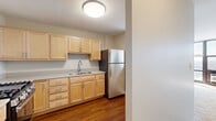 Located at 2701 S Indiana Ave in Chicago's South Side, this single-floor unit offers a clean, functional layout with a neutral color palette throughout. The entryway greets residents with a solid white door featuring classic brass hardware, opening into a corridor finished with rich hardwood-style flooring that extends through the main living and dining areas.

The hallway is flanked by ample built-in closet storage, including multiple floor-to-ceiling white panel closets with generous rod and shelf space — a practical and organized storage solution. The corridor leads naturally into the main living area, which is finished with plush wall-to-wall carpeting and bathed in natural light through large floor-to-ceiling windows. These expansive windows frame elevated views of the surrounding Chicago cityscape and green treetops, creating a bright and open ambiance.

A dedicated dining or flex area adjacent to the living space features hardwood-style flooring and its own window, allowing additional natural light to flow through. The bedroom is generously sized with carpeted floors, wide windows, and a built-in closet system with bifold doors offering substantial storage capacity. Throughout the unit, the design is straightforward and practical, emphasizing light, storage, and elevated views of the city.