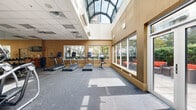 This fitness center offers a thoughtfully designed single-floor layout that provides ample space for a full range of exercise activities. The facility is divided into distinct workout zones, seamlessly connected under a combination of a suspended tile ceiling and a dramatic arched skylight that floods a portion of the space with abundant natural light.

The cardio zone is lined with multiple treadmills and elliptical machines, positioned along large floor-to-ceiling windows that offer views of a landscaped outdoor area, creating an open and energizing atmosphere during workouts. A stationary bike rounds out the cardio offerings. Adjacent to this area, a dedicated strength training section houses a multi-rack dumbbell station with a full range of free weights, an adjustable weight bench, and a cable/resistance machine, all arranged on durable rubber-flecked flooring designed for heavy use.

Full-length mirrors along one wall visually expand the space while serving a functional purpose for form monitoring. A wall-mounted television is positioned for visibility throughout the cardio section. The warm tan-toned walls and neutral flooring contribute to an energizing yet comfortable ambiance. A glass-panel door provides direct access to an outdoor terrace, further enhancing the connection to the outdoors. The facility is well-ventilated with clearly marked exits for safety.