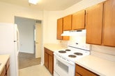 This single-floor unit offers a clean, functional layout across several well-defined living areas. The spacious living room is finished with neutral-toned wall-to-wall carpeting and features a large multi-pane window that fills the room with natural light and provides views of surrounding greenery, creating a bright and welcoming atmosphere. The kitchen is designed in a galley style, lined on both sides with warm honey-toned wood cabinetry offering ample storage. It is equipped with white appliances including a coil-top electric range with oven, an over-range ventilation hood, and a full-size refrigerator. Light-colored vinyl tile flooring and a double-basin sink with adjacent countertop workspace complete the practical setup. An opening at the end of the kitchen connects directly to the living area, promoting an open flow between spaces. The bedroom is a generously sized, carpet-floored room with white walls, a door with a brass-tone lever handle, and a floor-level heating vent. The full bathroom features warm beige tile flooring, a wood-cabinet vanity with a light countertop, a large wall mirror with overhead lighting, and a tub-shower combination equipped with safety grab bars and white tile surround. Overall, the unit presents a tidy, move-in-ready environment with consistent neutral finishes throughout.