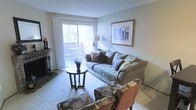 This single-floor space centers around a well-appointed living room designed for comfort and functionality. The room features a stone-faced fireplace along one wall, adding a warm and inviting focal point to the space. Soft, neutral-toned carpet covers the floor, complementing the light gray walls and creating a cohesive, understated aesthetic throughout.

Natural light fills the room through a large sliding glass door fitted with sheer white curtains, which opens to a private balcony where outdoor seating is visible. This connection to the outdoors enhances the sense of openness in the space. A textured ceiling adds subtle architectural detail overhead.

The living area is furnished with a generously sized tufted sofa accented with decorative pillows, paired with a patterned armchair and matching ottoman that introduce visual interest. A round pedestal side table anchors the seating arrangement, while a floor lamp and framed wall art contribute to the room's polished, livable character. Overall, the space presents a balanced blend of comfort, natural light, and classic residential design.