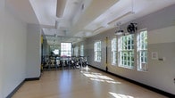 LC Idlewild's fitness studio is a well-appointed, single-floor exercise space designed to support a variety of workout routines. The room is defined by its bright, airy atmosphere, with a row of large, multi-pane windows along the walls that allow abundant natural light to pour in and offer views of the surrounding greenery outdoors. The light hardwood flooring reflects the sunlight beautifully, contributing to an open and energizing environment. A full-length mirror along one wall expands the sense of space and is a practical feature for form-checking during exercise. The studio is equipped with a fleet of stationary cycling bikes, making it suitable for group spin classes. TRX suspension training straps are mounted along the walls, enabling functional strength and bodyweight training. Ceiling-mounted fans ensure proper air circulation throughout the space, while a projector mounted from the ceiling suggests the room can support instructor-led or multimedia fitness sessions. The ceiling features a distinctive coffered design with structural beams, adding an architectural character that elevates the overall aesthetic of the fitness space. This studio is a thoughtfully designed, multi-use fitness facility built to accommodate both group classes and individual training needs.