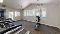 This single-floor fitness center at 9750 Old Placerville Rd in Sacramento, California offers a well-equipped space designed to support a variety of workout routines. The facility features vaulted ceilings that lend an open, airy feel throughout, complemented by multiple ceiling fans that promote air circulation during workouts. Large windows line the walls, flooding the interior with generous natural light and providing views of the surrounding greenery outside.

The main workout area is outfitted with a selection of True-brand cardio machines, including multiple treadmills, an elliptical trainer, and a stationary upright bike, allowing users to engage in diverse cardio exercises. Wall-mounted flat-screen televisions are positioned for easy viewing from the equipment. A dedicated open stretching zone features yoga mats on the floor, offering space for warm-ups, cool-downs, or mat-based exercises.

An additional room within the facility provides access to strength and resistance training equipment, expanding the range of available workouts. The flooring throughout consists of durable, patterned carpet tiles suited for a fitness environment. Entry to the facility is through glass-paneled doors, and the space is noted to be under video surveillance. The overall atmosphere is clean, functional, and bright, making it a practical and accessible fitness resource.