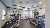 This fitness center is a thoughtfully designed, single-floor facility built to support a well-rounded workout routine. The space features a distinctive tray ceiling accented with dark crown molding and equipped with a large, modern ceiling fan, contributing both visual elegance and effective air circulation throughout the room. Recessed lighting provides consistent, bright illumination across the space.

The rubber flooring — speckled in a dark, durable finish — covers the entire workout area, providing cushioning and stability for all types of exercise. A multi-station cable strength machine serves as a centerpiece for resistance training, complemented by a recumbent bike, an upright stationary bike, a treadmill, an elliptical, and rowing machines, offering comprehensive cardio and strength options.

Wall-mounted mirrors in the cardio section enhance the sense of space and allow users to monitor their form during exercise. Natural light filters in through multiple windows fitted with horizontal blinds, keeping the environment bright and inviting. A built-in water fountain is conveniently located near the entrance for easy hydration access. The facility also includes a dedicated storage cubby unit and connects through glass-paneled doors to an adjacent outdoor area, allowing for seamless indoor-outdoor access.