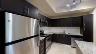 This contemporary single-floor space is thoughtfully designed with a clean, modern aesthetic throughout. The kitchen serves as a standout feature, boasting a full suite of stainless steel appliances — including a full-size refrigerator, electric range, built-in microwave, and dishwasher. Rich, dark espresso cabinetry lines the walls from floor to ceiling, offering generous storage and a bold visual contrast against the light granite countertops. A classic subway tile backsplash adds a refined touch, while the undermount sink with a modern faucet provides both style and functionality. The L-shaped kitchen layout maximizes workspace and traffic flow, making it as practical as it is attractive. Recessed tray ceiling detailing adds architectural interest and depth to the space, while track lighting ensures the kitchen is well-lit for both cooking and entertaining. Light-toned tile flooring ties the space together with a neutral, versatile foundation. Overall, the space presents a polished and functional environment with a cohesive modern design language, making it well-suited for those who appreciate contemporary style and thoughtful layout planning.