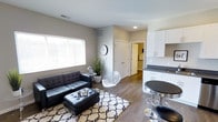 This 1-bedroom, 1-bathroom residence is thoughtfully designed to maximize every square foot, offering a cohesive and modern living experience across a single floor. The open-concept living and kitchen area serves as the heart of the home, where rich hardwood-style flooring and neutral gray walls create a clean, contemporary backdrop. The living area is bright and inviting, benefiting from a large window with horizontal blinds that allows ample natural light to fill the space. The kitchen features crisp white cabinetry, dark countertops, and an efficient layout that opens seamlessly to the living area, complemented by a round bistro-style dining table and bar seating — ideal for casual meals. A hallway connects the main living space to the bedroom and bathroom, maintaining a clear and functional flow throughout the unit. Recessed lighting and ceiling-mounted vents are integrated discreetly into the design, contributing to a polished, well-finished aesthetic. The overall ambiance is modern and streamlined, with a monochromatic color palette that lends itself to versatile styling. This unit delivers a compact yet comfortable living environment with thoughtful design details at every turn.