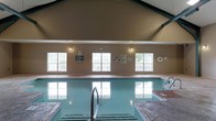 This single-floor indoor pool facility on Hearthside Drive in Wilmington, North Carolina, offers a well-appointed aquatic environment designed for both recreation and relaxation. The centerpiece of the space is a generously sized indoor swimming pool, clearly marked with depth indicators, and equipped with stainless steel entry rails for safe and easy access. Adjacent to the pool is a hot tub, providing a dedicated space for hydrotherapy and leisure. The room is crowned by a striking vaulted ceiling with a distinctive ridge, lending the space an open, airy feel. Natural light floods the interior through a row of large, multi-pane windows along one wall, offering views of the surrounding greenery while maintaining a bright and welcoming atmosphere. The walls feature a warm neutral paneling that complements the overall aesthetic. Safety signage and a life preserver ring are prominently displayed, reflecting a commitment to guest well-being. The pool deck features tiled surrounds, and the clean, well-maintained environment makes this facility a practical and enjoyable amenity for all users.