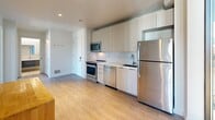 Located at 5522 Baum Blvd in Pittsburgh, PA, this efficiently designed 2-bedroom, 1-bathroom unit offers a modern, contemporary aesthetic throughout. The open-concept kitchen and living area serves as the heart of the space, featuring light wood-tone cabinetry extending to the ceiling, stainless steel appliances including a refrigerator, dishwasher, range, and microwave, complemented by clean white countertops and light hardwood-style flooring. A floor-to-ceiling glass sliding door floods the area with natural light and provides access to outdoor views. A compact bar-height dining table with stools adds a functional dining element to the space. The bedrooms are thoughtfully appointed, with the primary bedroom featuring floor-to-ceiling windows that frame views of the Pittsburgh streetscape, a built-in Murphy bed with integrated shelving, and a closet. The den also offers large windows with abundant natural light. The bathroom is finished with neutral tile, a walk-in glass-enclosed shower with a rainfall showerhead, mosaic tile flooring, and a floating vanity. A dedicated utility room rounds out the layout, contributing to the unit's practical and well-organized design.