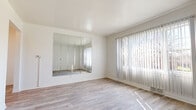 This single-floor unit offers a clean, functional layout with a cohesive design throughout. Light wood-style flooring flows seamlessly across the main living areas and bedrooms, creating a warm and unified aesthetic. The spacious living room is a standout feature, highlighted by a large wall-mounted mirror panel that amplifies the sense of space, paired with floor-to-ceiling vertical blinds that filter abundant natural light from an oversized window with outdoor views. An additional room with a window and horizontal blinds provides flexibility as a bedroom or secondary living space. The kitchen is well-equipped with light wood cabinetry, granite-patterned countertops, a gas range, dishwasher, and tile flooring laid in a diagonal pattern, offering ample storage and counter workspace. The bathroom features white tile wall surrounds, a tub-shower combination, a vanity with a mirror, a window for natural ventilation, and durable tile flooring. Throughout the unit, crisp white walls and clean baseboards maintain a bright, airy atmosphere. The thoughtful layout, consistent finishes, and generous natural light make this a highly functional and inviting space.