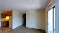 This single-floor space presents a straightforward and functional layout. The main room is finished with wall-to-wall carpeting in a neutral tone, providing a clean and understated foundation. The walls are painted in a light, off-white color, creating a bright and open atmosphere. A textured popcorn ceiling adds a classic architectural detail overhead, and a centrally located ceiling vent ensures efficient climate control throughout the space. Dark wood baseboards run along the perimeter of the room, offering a subtle contrast to the lighter walls and flooring. Multiple electrical outlets are positioned along the walls, adding practical convenience. A doorway at the far end of the room opens to an adjoining area, hinting at a connected layout beyond. The overall ambiance of the space is calm and neutral, offering a blank canvas that is well-suited for a variety of uses. The simplicity of the design, combined with its functional features, makes it an adaptable and versatile environment.