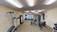 This fitness center is designed to support a well-rounded workout routine within a comfortably sized, single-level space. The room features neutral beige walls, white trim, and durable patterned carpet flooring throughout, creating a clean and functional environment. Multiple large, flush-mounted fluorescent light panels illuminate the space evenly, complemented by natural light streaming through several windows dressed with white shutters, including an arched accent window that adds a subtle architectural detail.

The cardio area is positioned along the windowed wall and includes two treadmills and an elliptical trainer, along with a wall-mounted television for entertainment during workouts. Strength training is well represented with a multi-station cable machine, a power tower for bodyweight exercises, and a flat weight bench. A two-tier dumbbell rack stocked with a range of free weights provides additional versatility for resistance training. A full-length mirror is mounted near the entrance, useful for monitoring form, and a stability ball rounds out the functional fitness accessories available.

A cork bulletin board is mounted on the wall, likely used for community announcements or fitness resources. The layout is open and uncluttered, allowing for ease of movement between equipment zones. Overall, the space offers a practical and inviting setting suited to a variety of fitness needs.