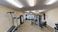 This fitness center is designed to support a well-rounded workout routine within a comfortably sized, single-level space. The room features neutral beige walls, white trim, and durable patterned carpet flooring throughout, creating a clean and functional environment. Multiple large, flush-mounted fluorescent light panels illuminate the space evenly, complemented by natural light streaming through several windows dressed with white shutters, including an arched accent window that adds a subtle architectural detail.

The cardio area is positioned along the windowed wall and includes two treadmills and an elliptical trainer, along with a wall-mounted television for entertainment during workouts. Strength training is well represented with a multi-station cable machine, a power tower for bodyweight exercises, and a flat weight bench. A two-tier dumbbell rack stocked with a range of free weights provides additional versatility for resistance training. A full-length mirror is mounted near the entrance, useful for monitoring form, and a stability ball rounds out the functional fitness accessories available.

A cork bulletin board is mounted on the wall, likely used for community announcements or fitness resources. The layout is open and uncluttered, allowing for ease of movement between equipment zones. Overall, the space offers a practical and inviting setting suited to a variety of fitness needs.