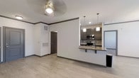 This single-story space showcases a thoughtfully designed open-concept layout that seamlessly connects the living area to the kitchen. Light hardwood-style flooring runs throughout, complementing the crisp white walls and dark baseboards with crown molding detail. A ceiling fan with a light fixture overhead serves the living area, providing both airflow and illumination.

The kitchen is a standout feature, equipped with dark gray cabinetry offering generous storage, a granite-look countertop peninsula that doubles as a breakfast bar with seating. Stainless steel appliances, including a range, microwave, and refrigerator, are neatly integrated into the design. A subway tile backsplash adds a modern touch, while two pendant lights suspended above the peninsula provide focused task lighting.

A doorway visible from the main living space leads to additional areas of the unit, and a large return air vent is prominently installed on one wall, reflecting practical HVAC considerations. The overall ambiance is clean, modern, and functional, with neutral tones and consistent finishes creating a cohesive aesthetic throughout the space.