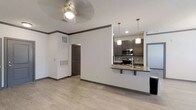 This single-story space showcases a thoughtfully designed open-concept layout that seamlessly connects the living area to the kitchen. Light hardwood-style flooring runs throughout, complementing the crisp white walls and dark baseboards with crown molding detail. A ceiling fan with a light fixture overhead serves the living area, providing both airflow and illumination.

The kitchen is a standout feature, equipped with dark gray cabinetry offering generous storage, a granite-look countertop peninsula that doubles as a breakfast bar with seating. Stainless steel appliances, including a range, microwave, and refrigerator, are neatly integrated into the design. A subway tile backsplash adds a modern touch, while two pendant lights suspended above the peninsula provide focused task lighting.

A doorway visible from the main living space leads to additional areas of the unit, and a large return air vent is prominently installed on one wall, reflecting practical HVAC considerations. The overall ambiance is clean, modern, and functional, with neutral tones and consistent finishes creating a cohesive aesthetic throughout the space.