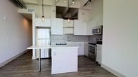 This single-floor space showcases a thoughtfully designed open-concept layout with a strong contemporary aesthetic. The kitchen is a standout feature, boasting soaring ceilings that amplify the sense of space and volume. Crisp white cabinetry lines the walls, complemented by a classic subway tile backsplash and gleaming white countertops that lend a clean, modern feel. Stainless steel appliances — including a full-size refrigerator, electric range, microwave, and dishwasher — add a polished, functional touch. A central kitchen island with a waterfall-edge countertop provides additional workspace and serves as a natural gathering point. Warm-toned hardwood flooring flows throughout, grounding the bright cabinetry and stainless finishes with an inviting richness. Industrial-style pendant lights with cage detailing hang above the island, while overhead track lighting ensures the space is well-lit and practical. Exposed ductwork near the ceiling reinforces the industrial-modern character of the space. The open layout encourages a seamless flow between kitchen and living areas, creating an environment that is both highly functional and visually striking.