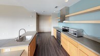 Located at 405 Bridge St NW in Grand Rapids, Michigan, this single-floor unit offers a thoughtfully designed layout with a blend of modern finishes and functional spaces. The open living area is anchored by rich, dark hardwood-style flooring and features expansive windows that frame views of the surrounding Grand Rapids skyline and cityscape, flooding the space with abundant natural light. A door provides access to an outdoor area, extending the living space further. The kitchen is well-equipped with light maple cabinetry, grey countertops, stainless steel appliances including a range and refrigerator, a stainless steel range hood, and open wood shelving accented by soft blue walls, creating a clean and contemporary cooking environment. A kitchen island with a sink adds additional prep space and functionality. The bedroom areas feature neutral grey carpeting and clean white walls, offering a calm and comfortable retreat. The bathroom is finished with dark hardwood-style flooring, a light maple vanity with grey countertop, and a backlit LED mirror, creating a bright and modern aesthetic. Throughout the unit, neutral tones, clean lines, and quality materials come together to create a cohesive and inviting living environment in the heart of Grand Rapids.