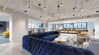 Located in Minneapolis, MN, the Malcolm Station 2nd Floor Community Room is a spacious, thoughtfully designed amenity space that blends modern aesthetics with comfortable, functional zones. The open-plan layout is anchored by a central lounge area featuring oversized tufted navy velvet sofas arranged around a patterned area rug, complemented by sculptural coffee tables. An artistic chandelier installation of bare Edison bulbs with draped black cords creates a dramatic focal point overhead.

A dedicated entertainment lounge offers bold yellow swivel chairs, round dark coffee tables, and a large wall-mounted TV set against a striking graphic mural, providing a vibrant, casual retreat. Nearby, a quiet seating nook with floor-to-ceiling windows frames outdoor views, bathing the space in natural light. A linear fireplace clad in slate tile anchors a separate lounge area with neutral seating.

The community kitchen and game zone features polished concrete floors, warm wood cabinetry with dark countertops, bar-height seating along a communal table, and a shuffleboard table. Exposed concrete columns and black-framed windows throughout lend an industrial-modern character, unifying the room's distinct areas into one cohesive, inviting environment.