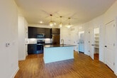 This well-appointed single-floor residence showcases a thoughtfully designed open-concept layout that seamlessly connects the kitchen and living areas. The kitchen serves as the centerpiece, featuring a large center island topped with dark granite countertops and an undermount sink fitted with a gold-toned faucet. Three glass pendant lights with brass fixtures hang above the island, providing both task lighting and visual elegance. Dark shaker-style cabinetry with gold hardware lines the kitchen walls, complemented by a herringbone-patterned tile backsplash and a full suite of stainless steel appliances, including a range, microwave, and refrigerator. Rich hardwood-style flooring flows throughout the main living areas, adding warmth and continuity. The living area opens to a private balcony through glass-paneled doors, allowing natural light to fill the space, while a ceiling fan ensures year-round comfort. The bedroom features plush carpeting, dual windows for natural light, and its own ceiling fan. The bathroom is finished with matching dark cabinetry with granite countertops, large-format tile flooring, and a glass-enclosed walk-in shower with black hardware accents. A dedicated laundry area and built-in shelving provide additional functionality and storage throughout this refined, move-in-ready home.